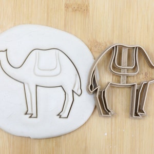 Può includere: Un tagliapasta in metallo a forma di cammello con una sella. Il tagliapasta è su una superficie di legno con un pezzo di pasta da modellare bianca dietro. Il tagliapasta è di qualità alimentare.
