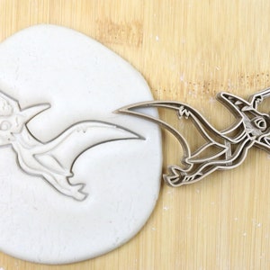 Könnte beinhalten: Ein Metall-Ausstecher in Form eines Pterodactylus-Dinosauriers. Der Ausstecher ist auf einer Holzoberfläche neben einem Stück weißen Teigs mit dem Abdruck des Ausstechers zu sehen.