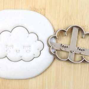 May include: A metal cookie cutter in the shape of a cloud with a face and eyelashes. The cutter is food grade and is shown on a wooden surface.