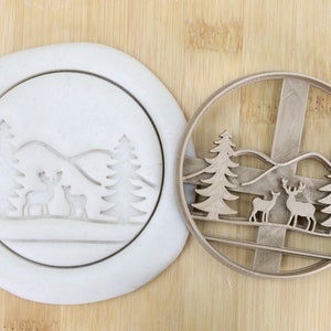 Puede incluir: Un cortador de galletas de plástico blanco con un diseño de dos ciervos de pie en un bosque nevado con árboles y colinas. El cortador de galletas es de calidad alimentaria y se muestra junto a una superficie de madera marrón.