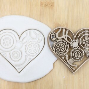 May include: A brown plastic cookie cutter in the shape of a heart with a steampunk design. The cutter is shown next to a white fondant heart with the same design.
