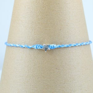 Könnte beinhalten: Ein blau-weiß geflochtenes Armband mit einem kleinen silbernen Sternanhänger. Das Armband wird vor einem neutralen Hintergrund präsentiert, der das filigrane Design und den Farbkontrast hervorhebt. Dieses Accessoire ist für den Alltag geeignet.