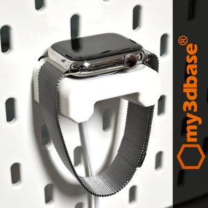 Op de afbeelding: Een witte 3D-geprinte wandbevestiging voor een Apple Watch met een zwarte Milanese Loop-band. Het horloge wordt opgeladen met een witte kabel. De houder is ontworpen om het horloge veilig vast te houden en gemakkelijk opladen mogelijk te maken.