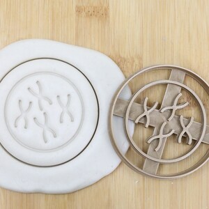 Könnte beinhalten: Ein brauner Plastik-Ausstecher in Form eines Kreises mit mehreren Chromosomenformen im Inneren. Der Ausstecher befindet sich auf einer Holzoberfläche mit einem weißen Stück Teig dahinter. Der Ausstecher ist mit "Food Grade" gekennzeichnet.