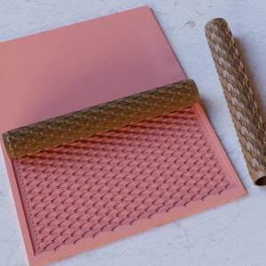 Könnte beinhalten: Ein brauner Nudelholz mit einem strukturierten Design rollt über eine rosa Silikonmatte mit dem gleichen Design. Die Matte liegt auf einer weißen Marmorfläche.