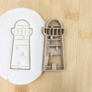 Può includere: Un tagliapasta in plastica marrone a forma di faro. Il tagliapasta è mostrato accanto a un'impronta di faro in pasta di zucchero bianca.