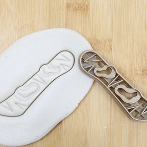 Könnte beinhalten: Ein hölzerner Ausstecher in Snowboardform und ein Abdruck in weißem Teig. Der Ausstecher hat ein detailliertes Design, und der Teig zeigt den Abdruck. Das Bild befindet sich auf einer hellen Holzoberfläche. Ein Lebensmittelqualitätssymbol befindet sich in der Ecke.