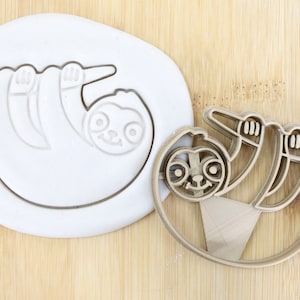 Puede incluir: Un cortador de galletas de plástico marrón en forma de perezoso colgado de una rama. El cortador está sobre una superficie blanca. El cortador es de calidad alimentaria.