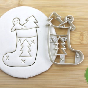 Puede incluir: Un cortador de galletas de pl&aacute;stico blanco en forma de calcet&iacute;n de Navidad con un hombre de jengibre y un &aacute;rbol de Navidad dentro. El cortador est&aacute; sobre una superficie de madera con una etiqueta "Food Grade" verde y blanca.