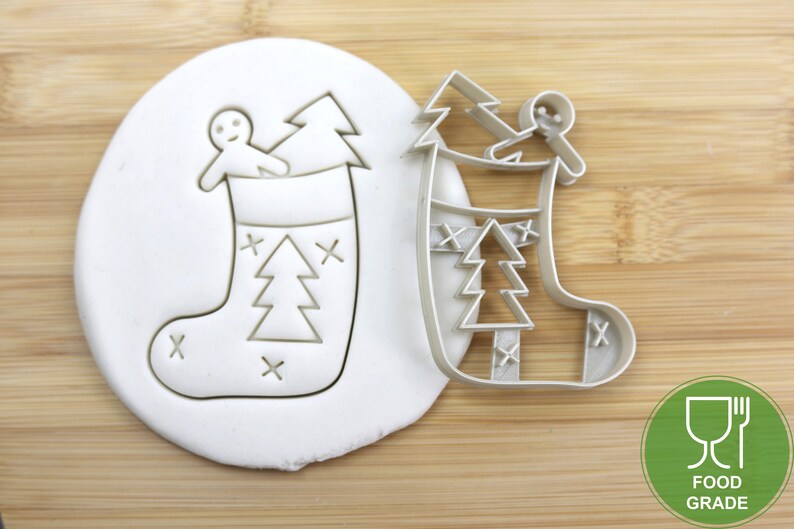 Puede incluir: Un cortador de galletas de pl&aacute;stico blanco en forma de calcet&iacute;n de Navidad con un hombre de jengibre y un &aacute;rbol de Navidad dentro. El cortador est&aacute; sobre una superficie de madera con una etiqueta "Food Grade" verde y blanca.