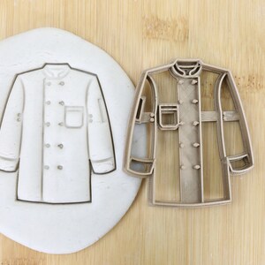 Puede incluir: Un cortador de galletas de plástico marrón en forma de chaqueta de chef. El cortador está sobre una superficie de madera junto a una impresión de fondant blanco del cortador.