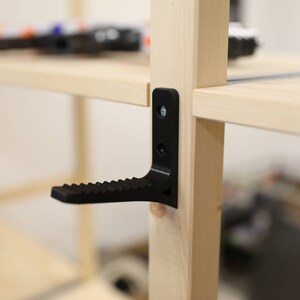 May include: A black plastic shelf support bracket with a step attached to the bottom. The bracket is attached to a wooden shelf unit.