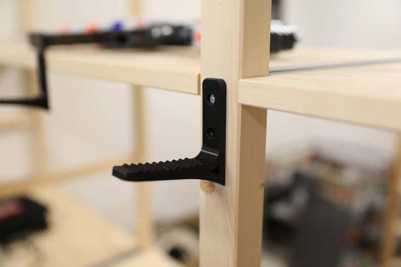 May include: A black plastic shelf support bracket with a step attached to the bottom. The bracket is attached to a wooden shelf unit.