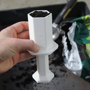 Puede incluir: Herramienta de jardinería de plástico blanco con una parte superior hexagonal llena de tierra. La herramienta tiene un mango y una base que también es hexagonal.