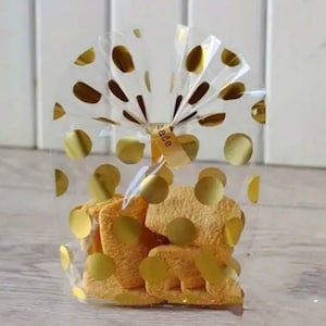 Puede incluir: Una bolsa de plástico transparente con lunares dorados y una cinta dorada atada en la parte superior. La bolsa contiene varias galletas pequeñas, doradas y cuadradas.
