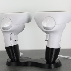 Könnte beinhalten: Ein schwarzer Plastikständer mit zwei weißen VR-Controllern. Die Controller haben schwarze Bänder und sind für die Verwendung mit einem Virtual-Reality-Headset konzipiert.