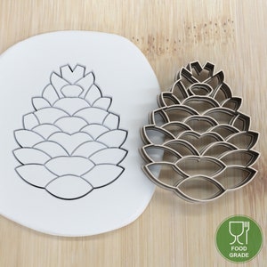 May include: A white plastic cookie cutter in the shape of a pine cone, next to a brown wooden cookie cutter in the same shape. Both cutters are food grade.