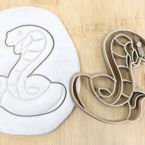 May include: A food-grade plastic cookie cutter in the shape of a coiled snake. The cutter is shown next to a piece of white modeling clay that has been pressed with the cutter.