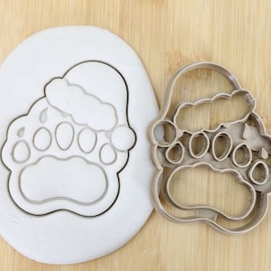 Könnte beinhalten: Ein brauner Plastik-Ausstecher in Form einer Bärenpfote, die einen Weihnachtsmann-Hut trägt. Der Ausstecher befindet sich auf einer weißen Oberfläche.