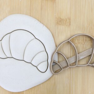 Könnte beinhalten: Ein brauner Plastik-Ausstecher in Form eines Croissants. Der Ausstecher liegt auf einer Holzoberfläche neben einem Stück weißen Teigs, in das die Umrisse eines Croissants geschnitten sind. Der Ausstecher ist mit "Food Grade" gekennzeichnet.