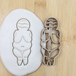 May include: A metal cookie cutter in the shape of a stylized female figure. The cutter is food grade and is shown next to a cookie dough imprint.