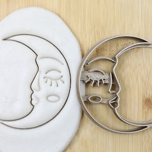 Pode incluir: Um cortador de biscoitos de metal prateado em forma de lua crescente com um rosto estilizado. O cortador é mostrado ao lado de uma impressão de argila branca do cortador.