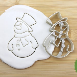 Puede incluir: Un cortador de galletas de metal plateado en forma de mu&ntilde;eco de nieve con un sombrero de copa negro y una bufanda. El cortador se muestra sobre una superficie de madera con una muestra de la masa de galletas cortada.