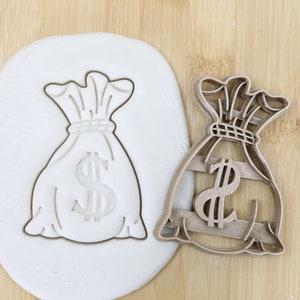 Könnte beinhalten: Ein brauner Plastik-Ausstecher in Form eines Geldbeutels mit einem Dollarzeichen darauf. Der Ausstecher befindet sich auf einer weißen Oberfläche mit braunem Holz Hintergrund. Der Ausstecher ist lebensmittelecht.