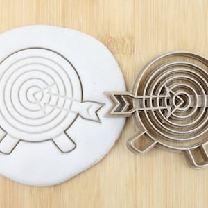 Puede incluir: Un cortador de galletas de madera con forma de diana con una flecha, junto a un círculo blanco de masa con la huella de la diana. El cortador tiene una etiqueta de calidad alimentaria.
