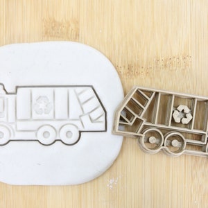 Może przedstawiać: Brązowa plastikowa foremka do ciasteczek w kształcie śmieciarki z symbolem recyklingu po bokach. Foremka do ciasteczek leży na drewnianej powierzchni obok białej silikonowej formy o tym samym wzorze.