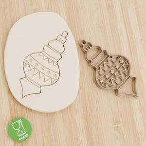 May include: A beige Christmas ornament cookie cutter and a cookie with the ornament design pressed into it. The cutter has a detailed design with stars and zigzags. A food-grade sticker is in the lower left corner.