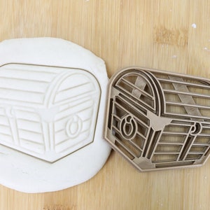 Könnte beinhalten: Ein goldfarbener Metall-Ausstecher in Form einer Schatztruhe. Der Ausstecher liegt auf einer Holzoberfläche mit einem weißen Fondant-Abdruck der Truhe.