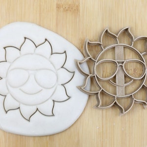 Puede incluir: Un cortador de galletas de color dorado en forma de sol con gafas de sol. El cortador está sobre una superficie de madera junto a una masa blanca con el mismo diseño impreso en ella. El cortador está etiquetado como "Food Grade".