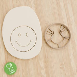 Könnte beinhalten: Ein beiger Smiley-Ausstecher neben einem Ausstechplätzchen mit einem Smiley-Design. Der Ausstecher ist hellbraun und hat einen Food Grade-Aufkleber. Die Artikel befinden sich auf einer Holzoberfläche.