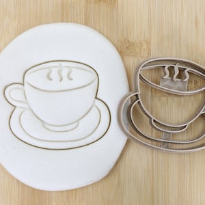 Könnte beinhalten: Ein Keksausstecher aus Metall in Form einer Kaffeetasse und Untertasse. Der Ausstecher ist lebensmittelecht und wird auf einer Holzoberfläche neben einem Stück weißen Modelliertones gezeigt.