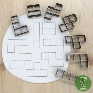 Könnte beinhalten: Ein Set mit 10 lebensmittelechten Keksausstechern in Form verschiedener Tetris-Blöcke. Die Ausstecher sind braun und auf einer weißen Oberfläche angeordnet. Der Text "FOOD GRADE" ist auf einem grünen Kreis mit einem Gabel- und Messer-Symbol gedruckt.