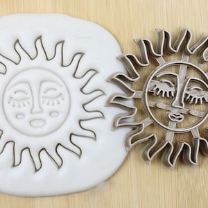 Op de afbeelding: Een metalen koekjesvormpje in de vorm van een zon met een gestileerd gezicht. De zon heeft gesloten ogen en een kleine mond. De koekjesvormpje is van voedselkwaliteit.