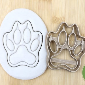 Puede incluir: Un cortador de galletas de metal plateado en forma de huella de pata. El cortador está sobre una superficie de madera. El cortador es de calidad alimentaria.