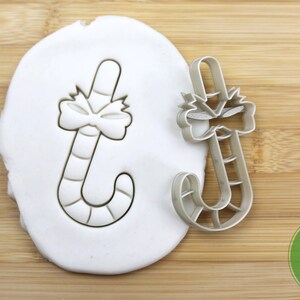 Puede incluir: Un cortador de galletas de color plateado en forma de bast&oacute;n de caramelo con un lazo en la parte superior. El cortador est&aacute; sobre una superficie blanca y un fondo de madera. El cortador es de calidad alimentaria.