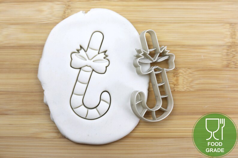 Puede incluir: Un cortador de galletas de color plateado en forma de bast&oacute;n de caramelo con un lazo en la parte superior. El cortador est&aacute; sobre una superficie blanca y un fondo de madera. El cortador es de calidad alimentaria.