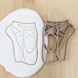 Könnte beinhalten: Ein brauner Plastik-Ausstecher in Form eines Paares Ballettschuhe. Der Ausstecher liegt auf einer Holzoberfläche neben einem Stück weißen Fondants, in das die Umrisse der Schuhe ausgeschnitten sind.