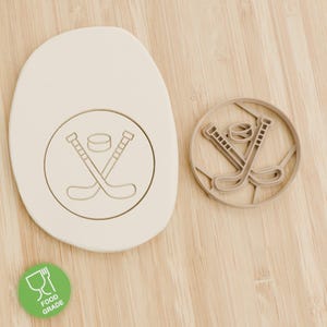 Könnte beinhalten: Ein beiger Hockey-Ausstecher und ein Keks mit dem Design von Hockeyschläger und Puck. Der Ausstecher ist rund und hat eine detaillierte Umrisszeichnung der Hockeyschläger und des Pucks. Ein grüner Aufkleber in Lebensmittelqualität befindet sich in der linken unteren Ecke.