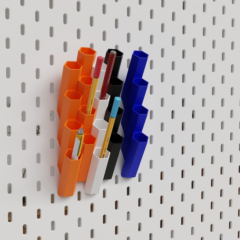 Puede incluir: Un conjunto de porta l&aacute;pices de pared de color naranja, blanco y azul con varios l&aacute;pices y bol&iacute;grafos de colores en su interior. Los porta l&aacute;pices est&aacute;n hechos de pl&aacute;stico y tienen forma hexagonal.