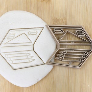 Könnte beinhalten: Ein Ausstecher aus Metall in Form eines Sechsecks mit einem Design aus Bleistiften, einem Dreieck und einem Würfel. Der Ausstecher liegt auf einem Stück weißen Teigs.