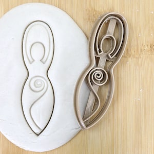 Könnte beinhalten: Ein brauner Plastik-Ausstecher in Form einer stilisierten weiblichen Figur mit einem Spiralmuster. Der Ausstecher liegt auf einer Holzoberfläche neben einem Stück weißen Teigs, aus dem das gleiche Muster ausgeschnitten ist. Der Text "FOOD GRADE" ist in Grün auf weißem Hintergrund sichtbar.