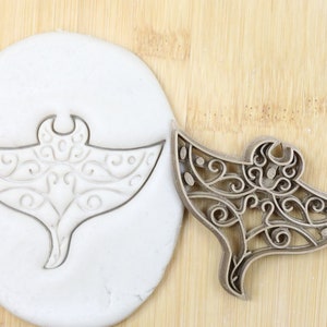 May include: A detailed, ornate, food-grade plastic cookie cutter in the shape of a manta ray. The cutter is shown pressed into a white fondant.