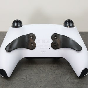 Könnte beinhalten: Ein weißer PlayStation 5 DualSense-Controller mit schwarzen Paddel-Anbauten auf der Rückseite. Der Controller hat ein schwarzes Sony-Logo auf der Vorderseite.