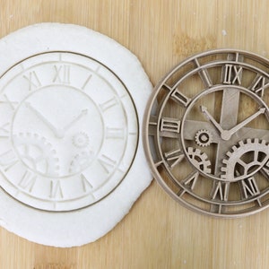 Pode incluir: Um cortador de biscoitos em forma de relógio e um biscoito com o desenho do relógio pressionado. O cortador é de cor castanho claro e apresenta algarismos romanos e engrenagens. O biscoito é branco e tem o mesmo desenho de relógio. A imagem está numa superfície de madeira.
