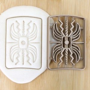 Puede incluir: Un cortador de galletas de plástico marrón en forma de escudo estilizado con un diseño geométrico. El cortador se muestra junto a una impresión blanca del diseño en masa.