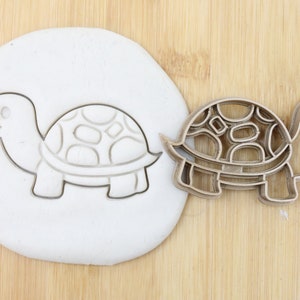 Puede incluir: Un cortador de galletas de metal en forma de tortuga con un diseño de caparazón detallado. El cortador se muestra junto a una pieza de arcilla de modelar blanca con una impresión del cortador.